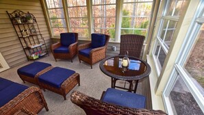 Un salón de sol con mobiliario de mimbre, una mesa de vidrio con una vela, y vistas a árboles a través de amplios ventanales.