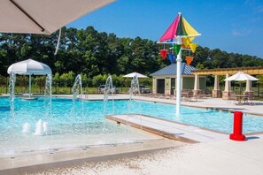 Una piscina con múltiples chorros de agua, un pabellón y una pérgola con sombrillas.