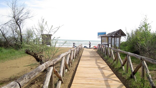 Un paseo marítimo de madera que lleva a una playa, con una pequeña construcción de madera y un letrero.