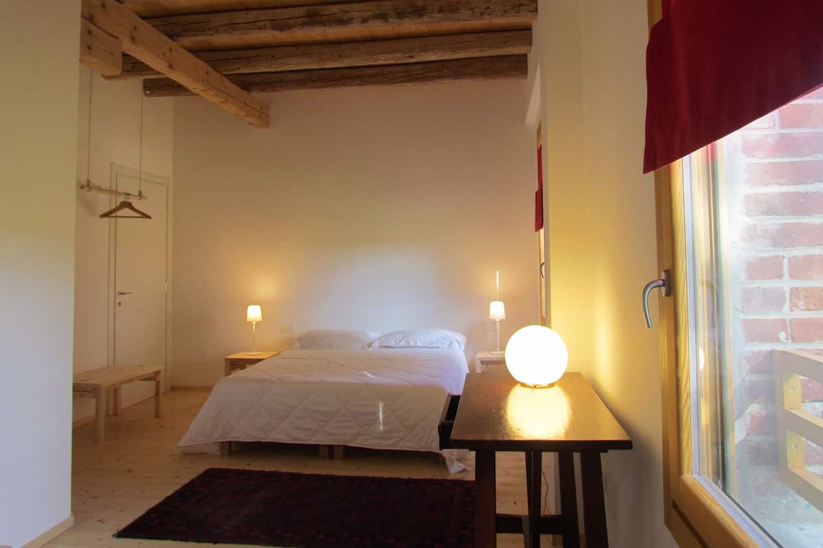 Ein Schlafzimmer mit einem Bett, Nachttischen, einer Hängelampe, einem Fenster mit Blick auf eine Ziegelwand und einer Holzbalkendecke.