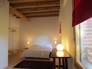 Un dormitorio con cama, mesitas de noche, una lámpara colgante, un ventanal con vista a un muro de ladrillo y techo de madera.