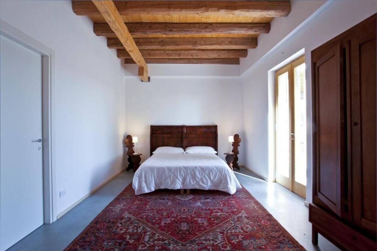Ein Schlafzimmer mit einem großen Bett, Holzbalkendecke, einem roten Teppich mit Muster und zwei Nachttischlampen.