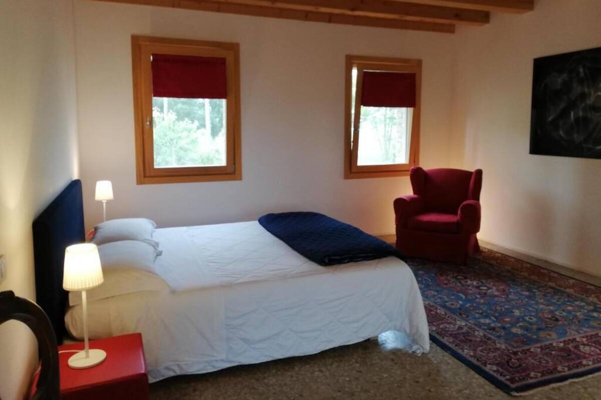 Ein Schlafzimmer mit einem Bett, zwei Fenstern mit roten Vorhängen, einem roten Sessel, einem Nachttisch mit Lampe und einem gemusterten Teppich.