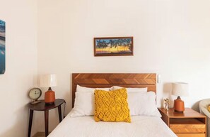 Een hotelkamer met een houten hoofdeinde, een bed met witte lakens, een geel gestreept kussen, twee nachtkastjes met lampen en een schilderij aan de wand.