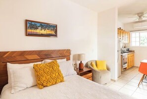 Ein Schlafzimmer mit einem Holzkopfende, einem Bett mit weißer Bettwäsche, einem gelben Zierkissen, einem Nachttisch mit Lampe und einem Palmenbaum-Gemälde an der Wand.