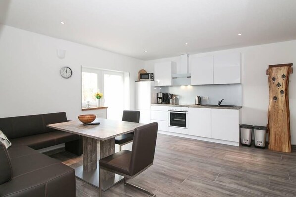 A modern kitchen with white cabinets, a microwave, and a coffee machine. There is a dining area with a wooden table and chairs.