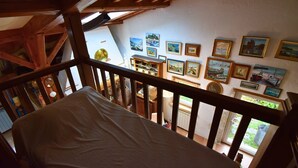 Een kamer met een houten plafond en een balkonhek. Er hangen verschillende afbeeldingen in lijstjes aan de muur.