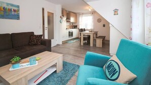 Un salon moderne avec un canapé, une table basse et un fauteuil bleu. On aperçoit un coin cuisine avec une table à manger et des chaises à travers une porte ouverte.