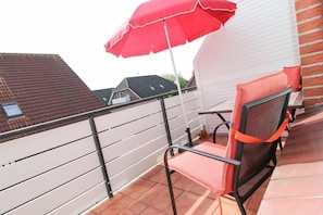 A balcony with a red umbrella, a chair, and a white wall.
