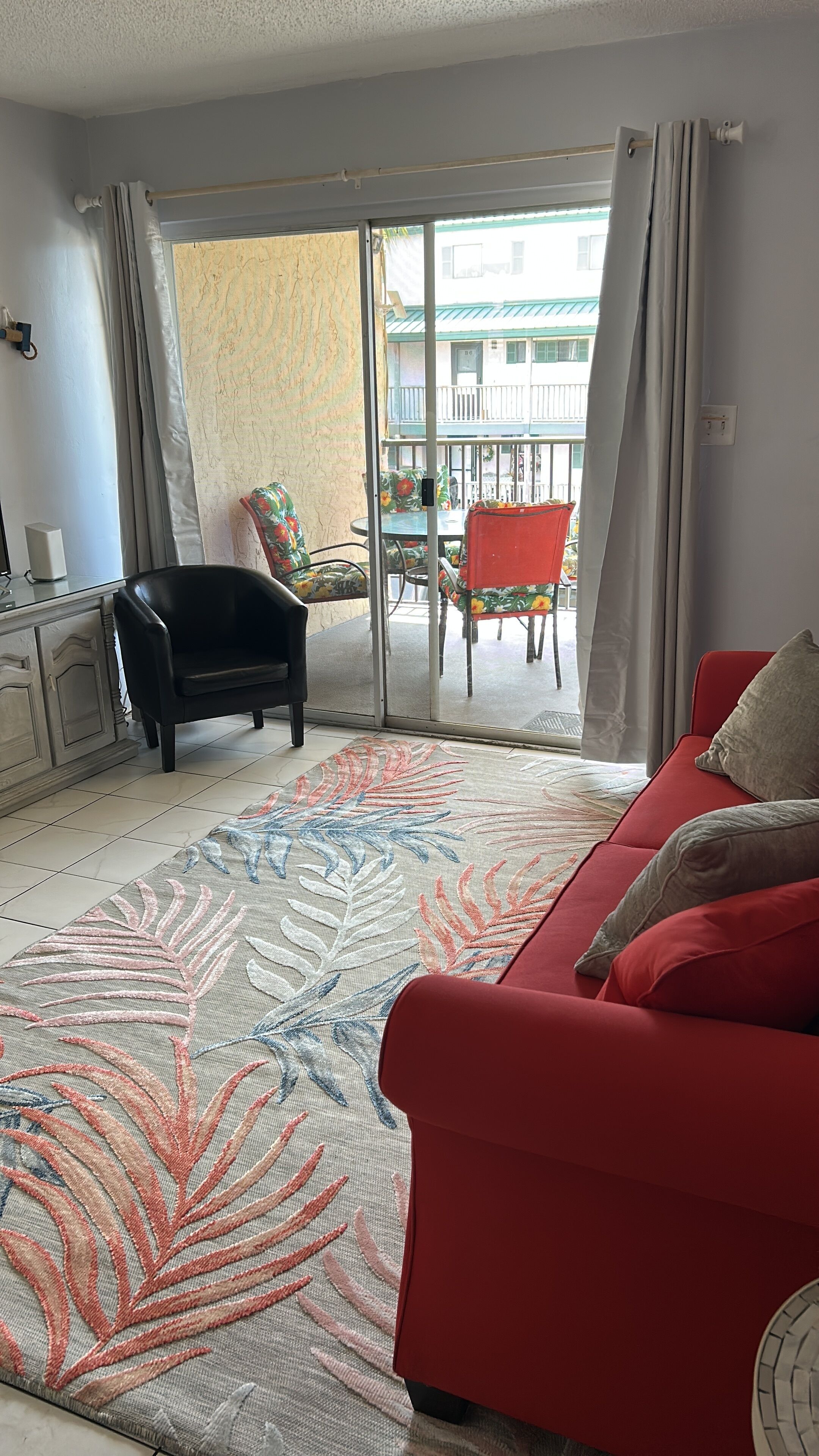 Ein Wohnzimmer mit einem roten Sofa, einem schwarzen Sessel, einem gemusterten Teppich und einer Schiebetür zu einem Außenbereich mit Tisch und Stühlen.