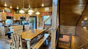 A rustic kitchen with wooden cabinets, a large wooden dining table, and a staircase with a wooden railing. - MORE THAN A GORGEOUS CABIN, IT'S AN EXPERIENCE!!!!  CLOSE TO ATTRACTIONS & GSM (Sevierville)