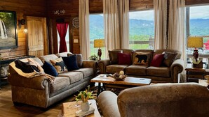 A cozy log cabin interior with a large sofa set, a coffee table, and a view of the mountains through large windows. - MORE THAN A GORGEOUS CABIN, IT'S AN EXPERIENCE!!!!  CLOSE TO ATTRACTIONS & GSM (Sevierville)