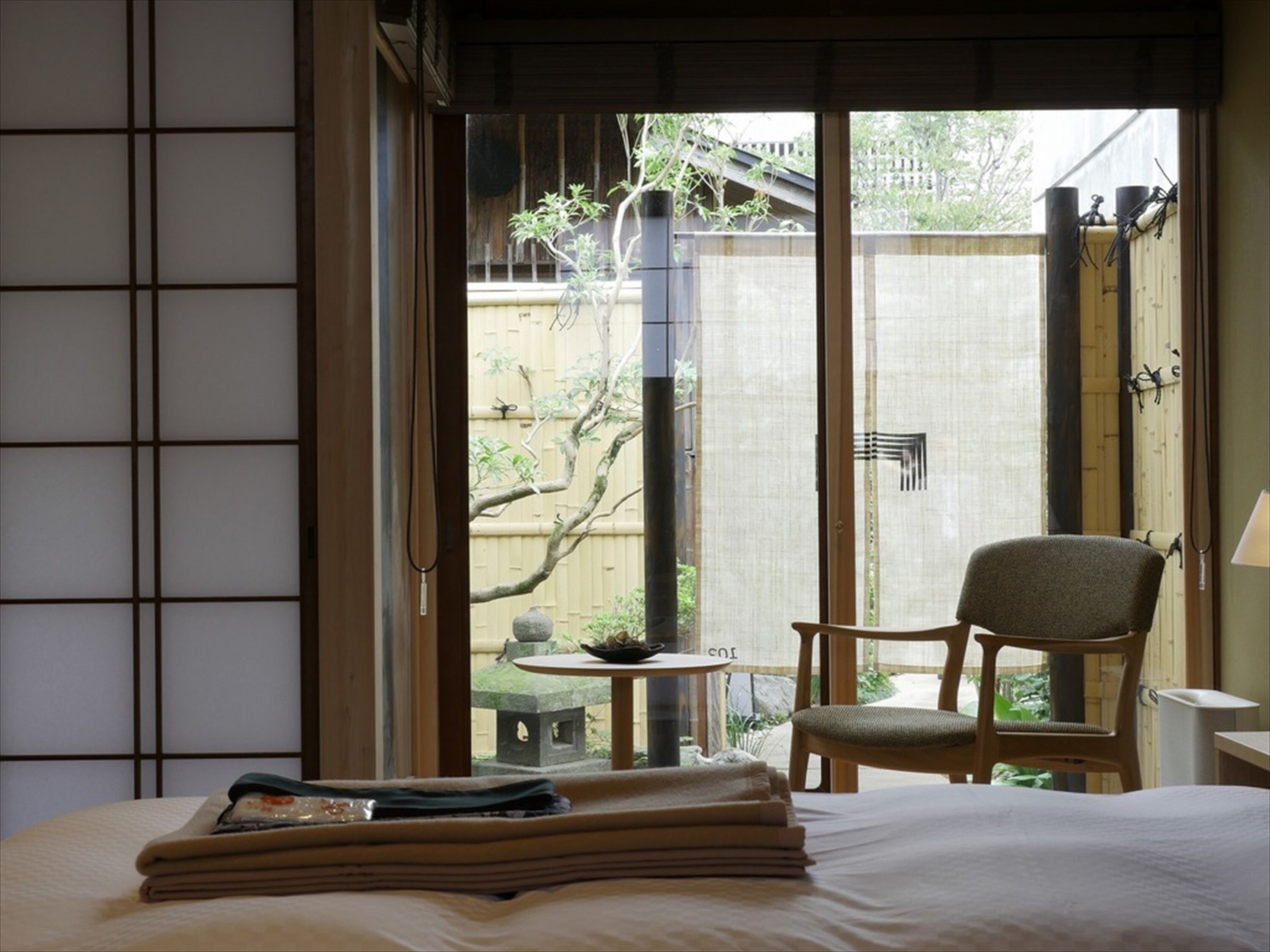 A traditional Japanese-style room with tatami flooring, a low table with a tea set, a chair, and a bed with neatly folded towels.