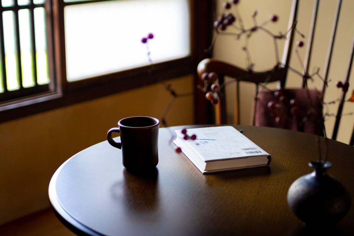 Una mesa con una taza negra y un libro, una silla y una ventana con barrotes.