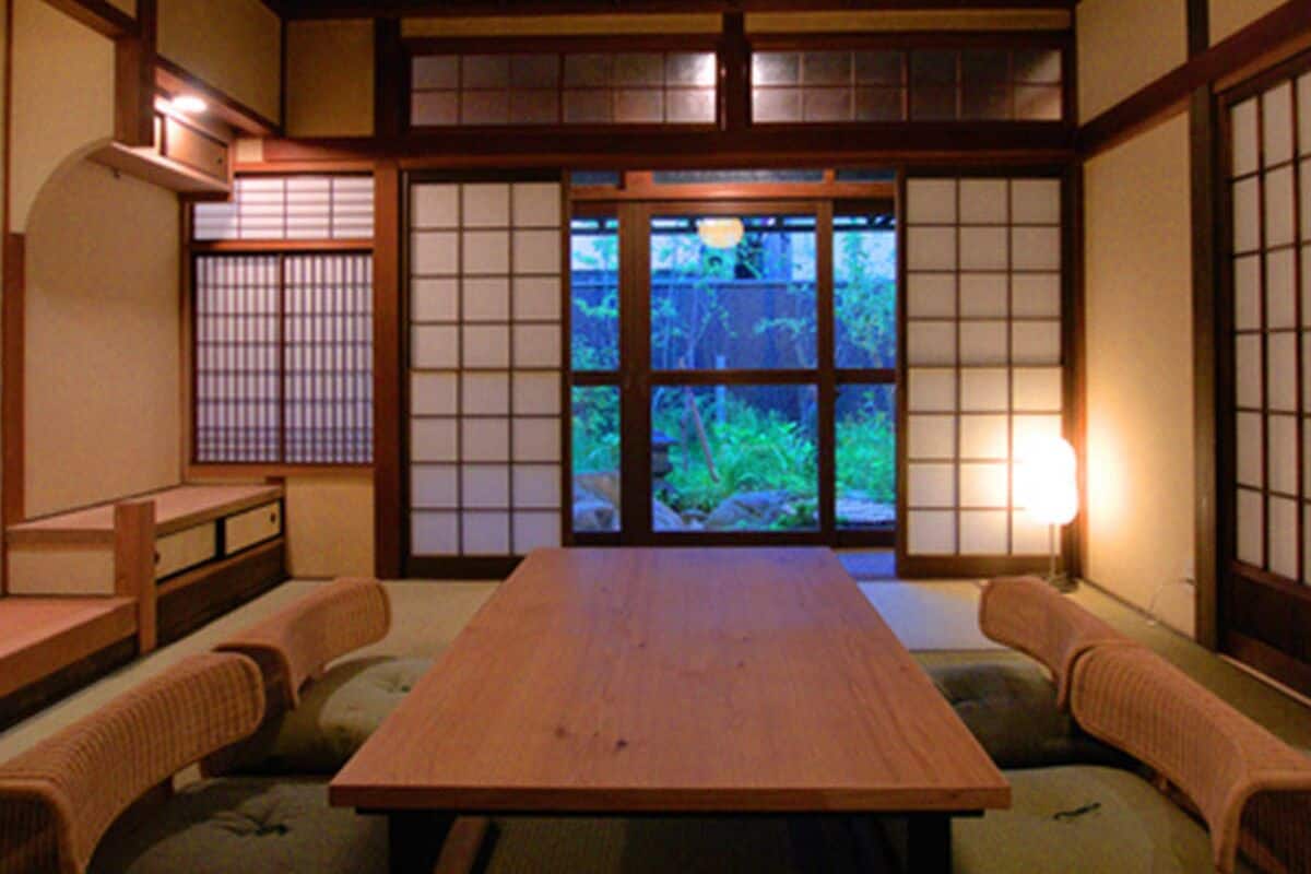 Una habitación tradicional japonesa con asientos de estera de tatami, una mesa de madera, puertas correderas y vistas a la vegetación.