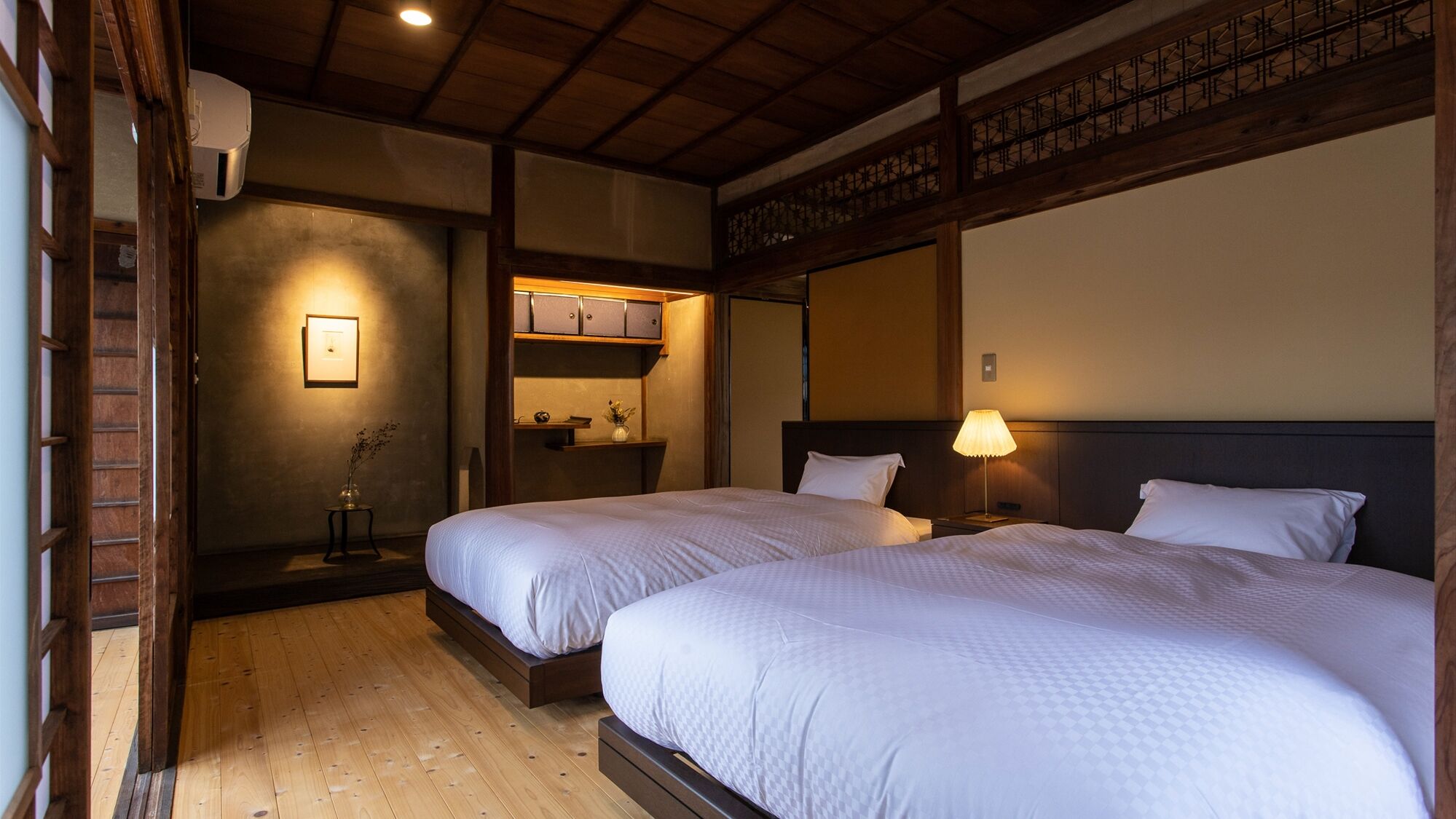 A hotel room with two beds, a wooden floor, a small table with a lamp, and a shelf with items.
