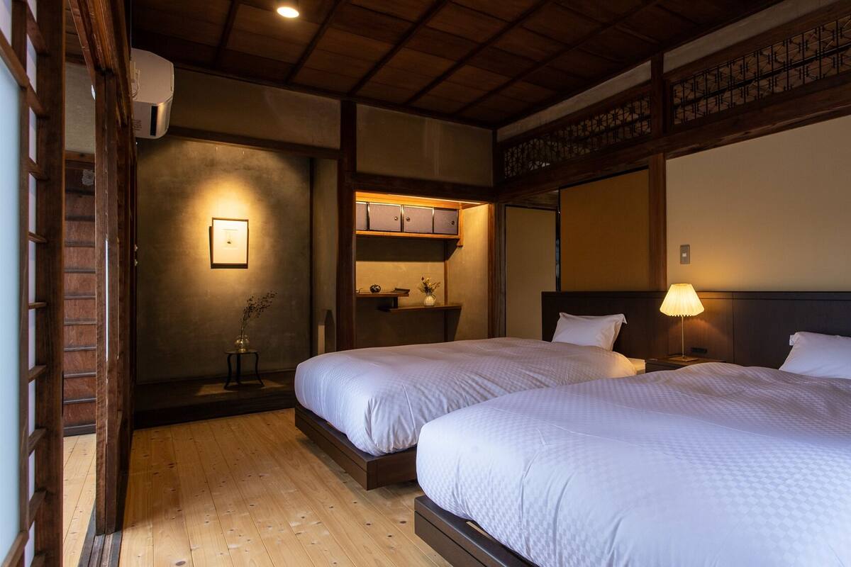 Habitación de hotel con dos camas, piso de madera, una mesita con lámpara y una estantería con objetos.