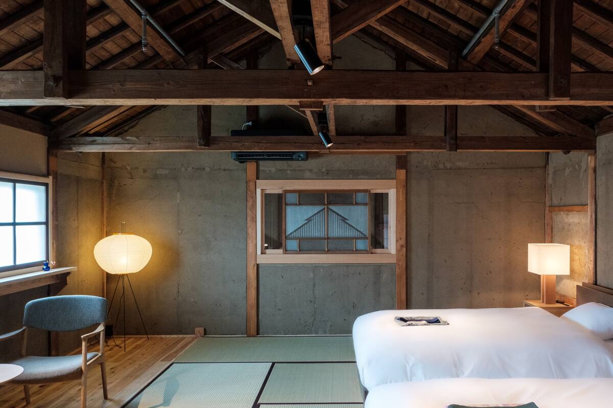 A room with wooden beams, tatami mats, a desk, two beds, and a window with a view of a bridge.