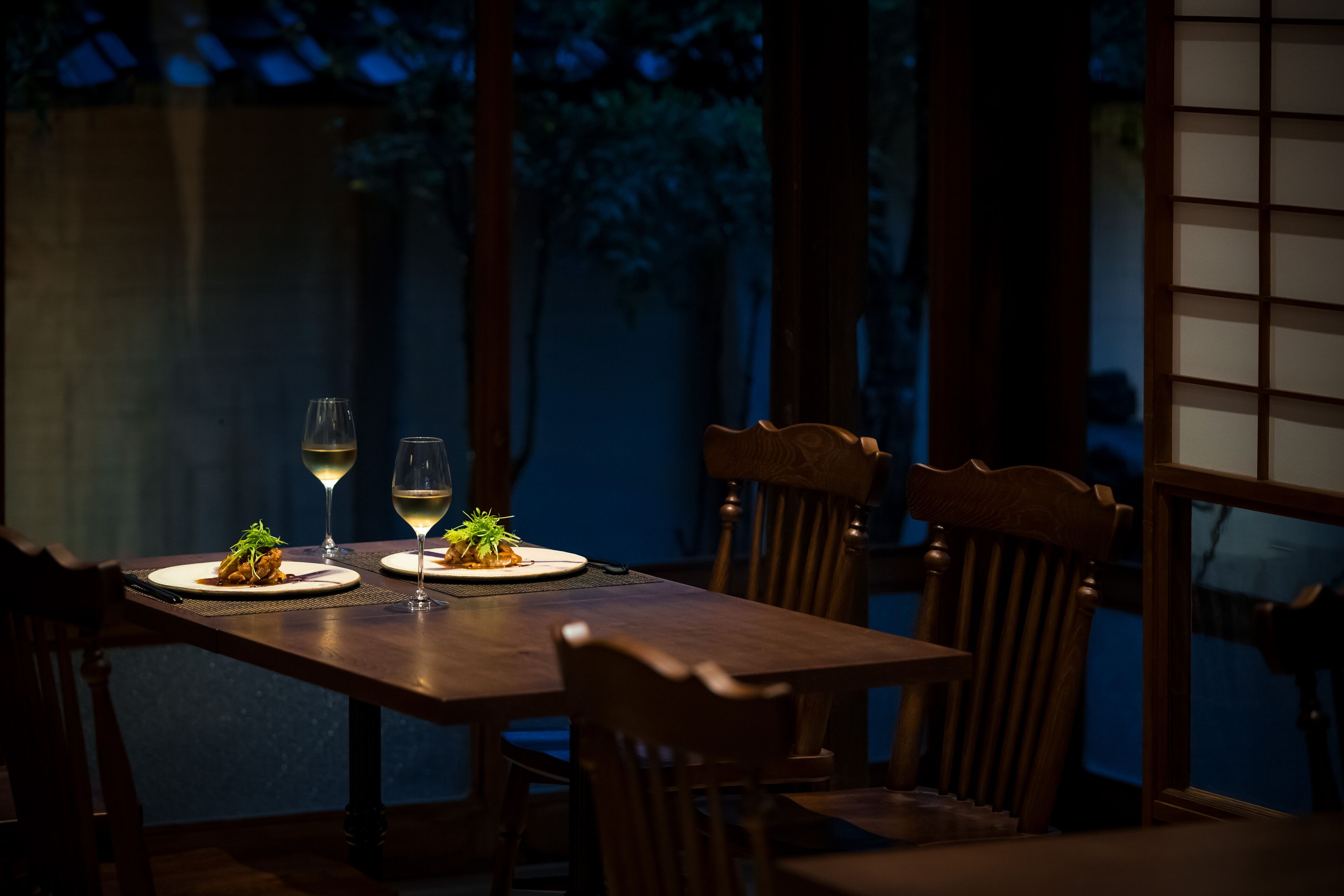 A dimly lit wooden dining table with two plates of food and two wine glasses, set against a backdrop of tall trees and a wooden structure.