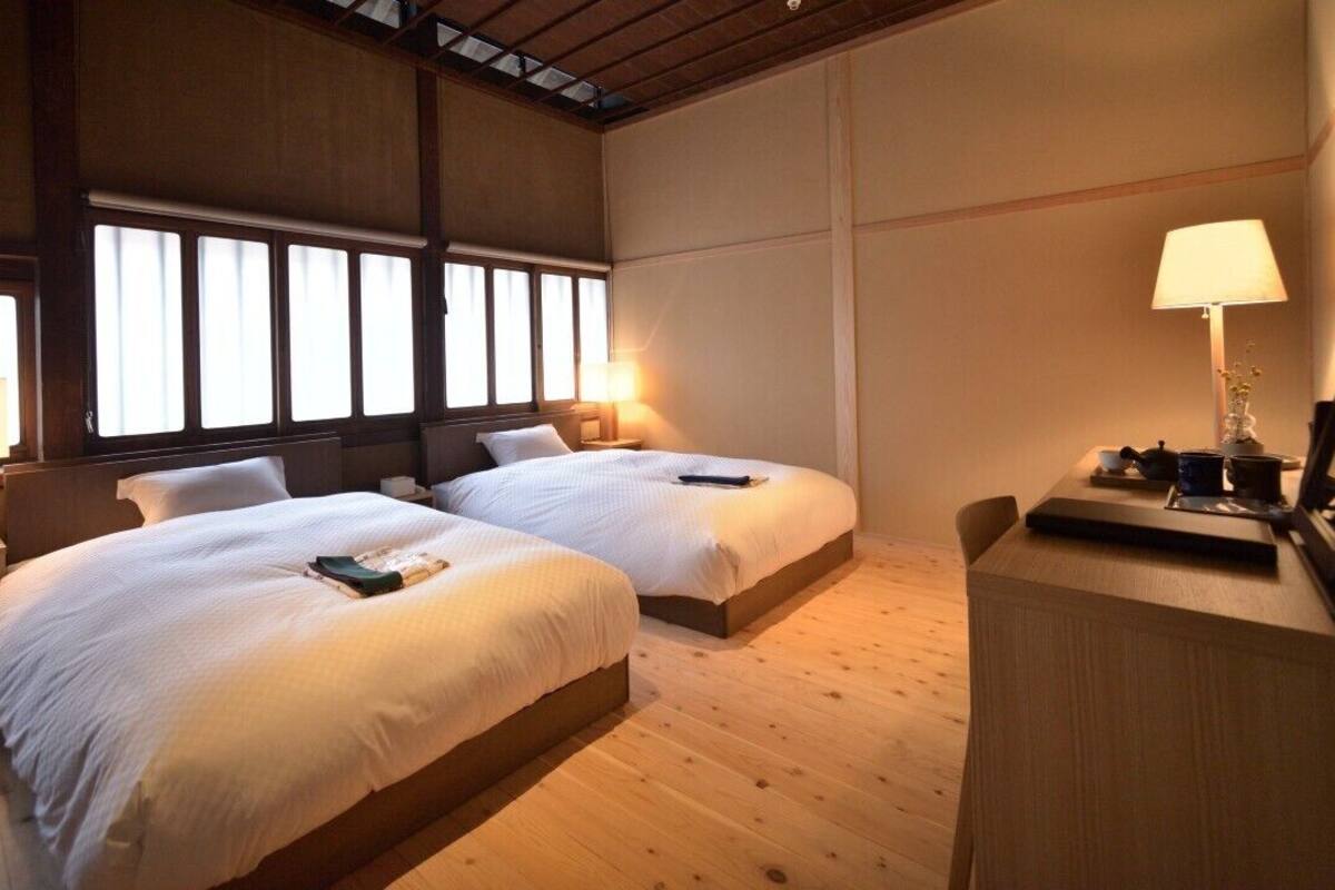 A room with two beds, wooden floors, a desk with a lamp, and a window with curtains.