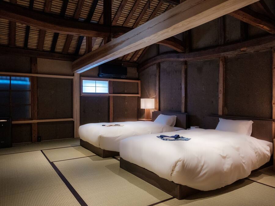 A traditional Japanese-style room with tatami flooring, two beds with white bedding, a wooden ceiling, and a small window.