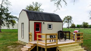 A small wooden structure with a red door, a small window, and a deck with steps leading to a table and chairs. - Queens Hotel- a cozy studio cottage in the country. Pool and hot tub.  (Round Top)