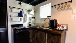 A kitchen with a black stove, wooden cabinets, a coffee maker, and a window above the sink. - Queens Hotel- a cozy studio cottage in the country. Pool and hot tub.  (Round Top)