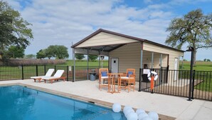A pool area with lounge chairs, a covered seating area, and a building with a door and windows. - Queens Hotel- a cozy studio cottage in the country. Pool and hot tub.  (Round Top)