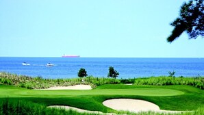 Un parcours de golf avec des bunkers et un mât de drapeau, offrant une vue sur une étendue d’eau où l’on aperçoit un navire au loin.