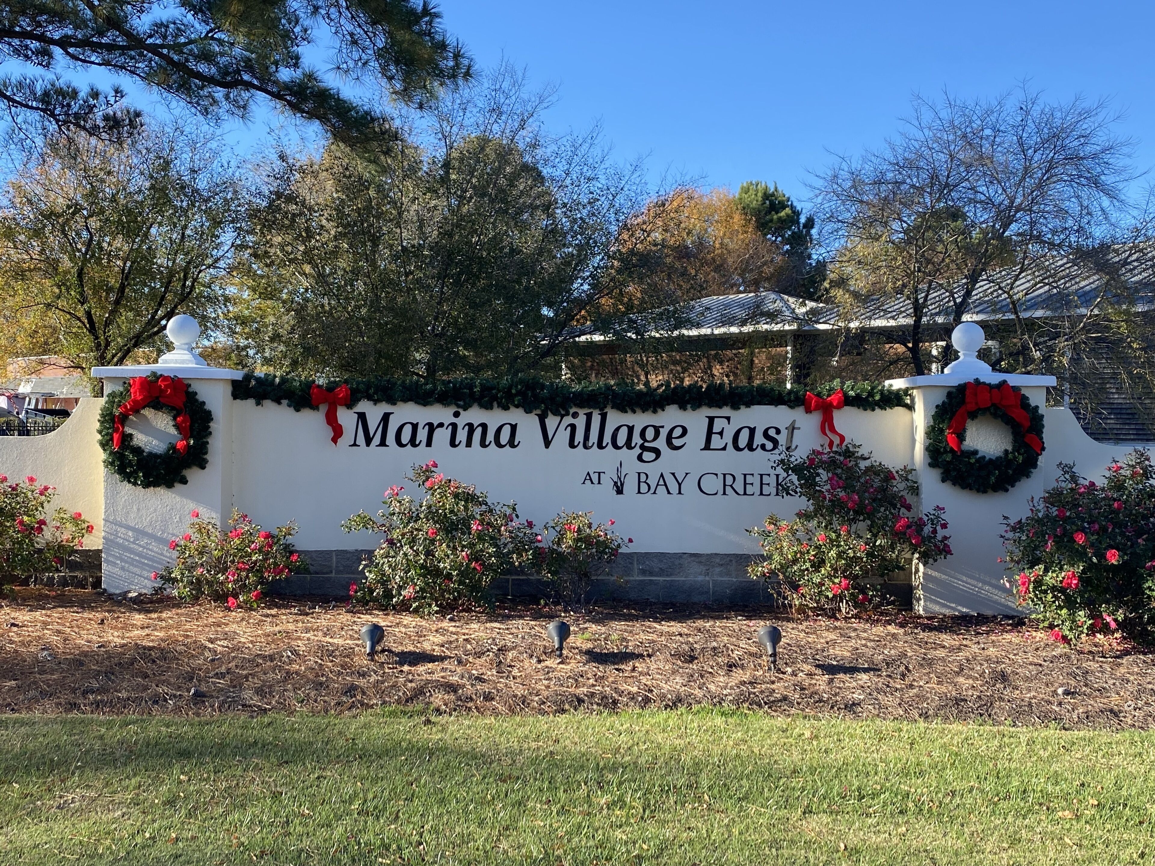 A sign for 'Marina Village East at Bay Creek' with wreaths and a clear blue sky in the background.