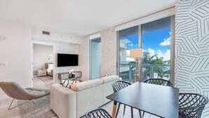 A modern living room with a sofa, a dining table, and chairs. There is a TV mounted on the wall, and a window with a view of palm trees. - Rare Modern 1Bdrm Condo- Miami Design Dist (Miami)