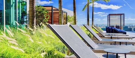 A rooftop lounge area with sun loungers and tables, palm trees, and a modern building with large windows.
