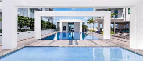 A modern pool area with clear blue water, surrounded by white buildings and palm trees.