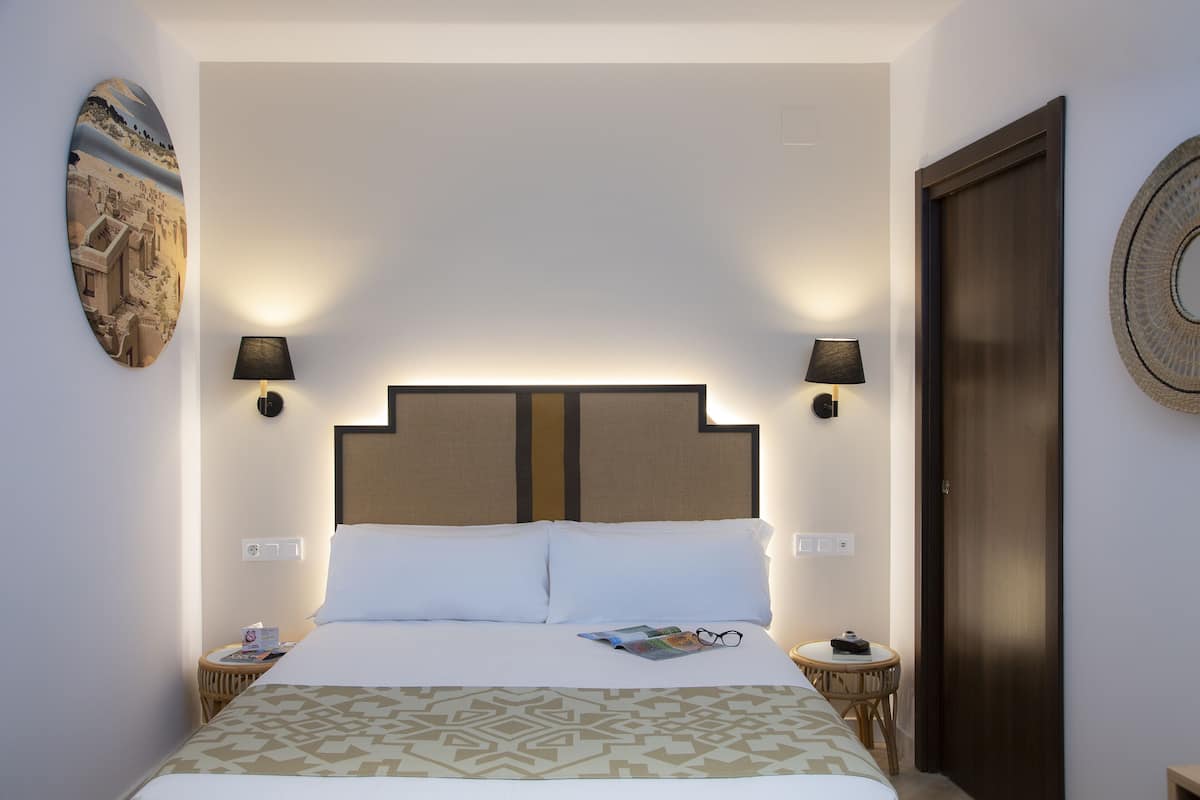 Een keurig opgemaakt bed met een beddekleed met patroon, twee nachtlampjes, een klein rond tafeltje met een boek en brillen, en een aan de muur hangend schilderij.