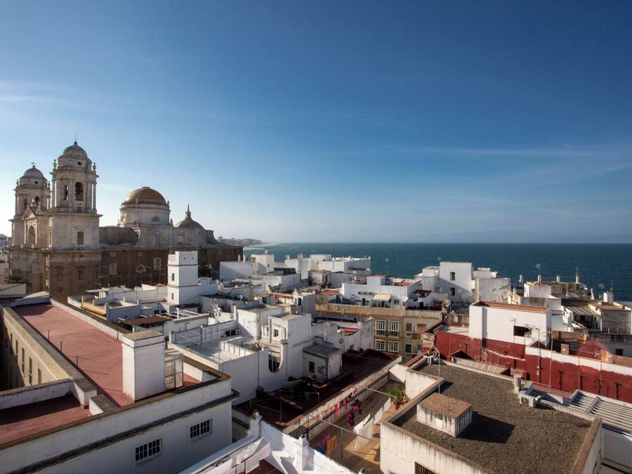 Un paisaje urbano costero con edificios blancos y una catedral destacada con dos torres y una cúpula.