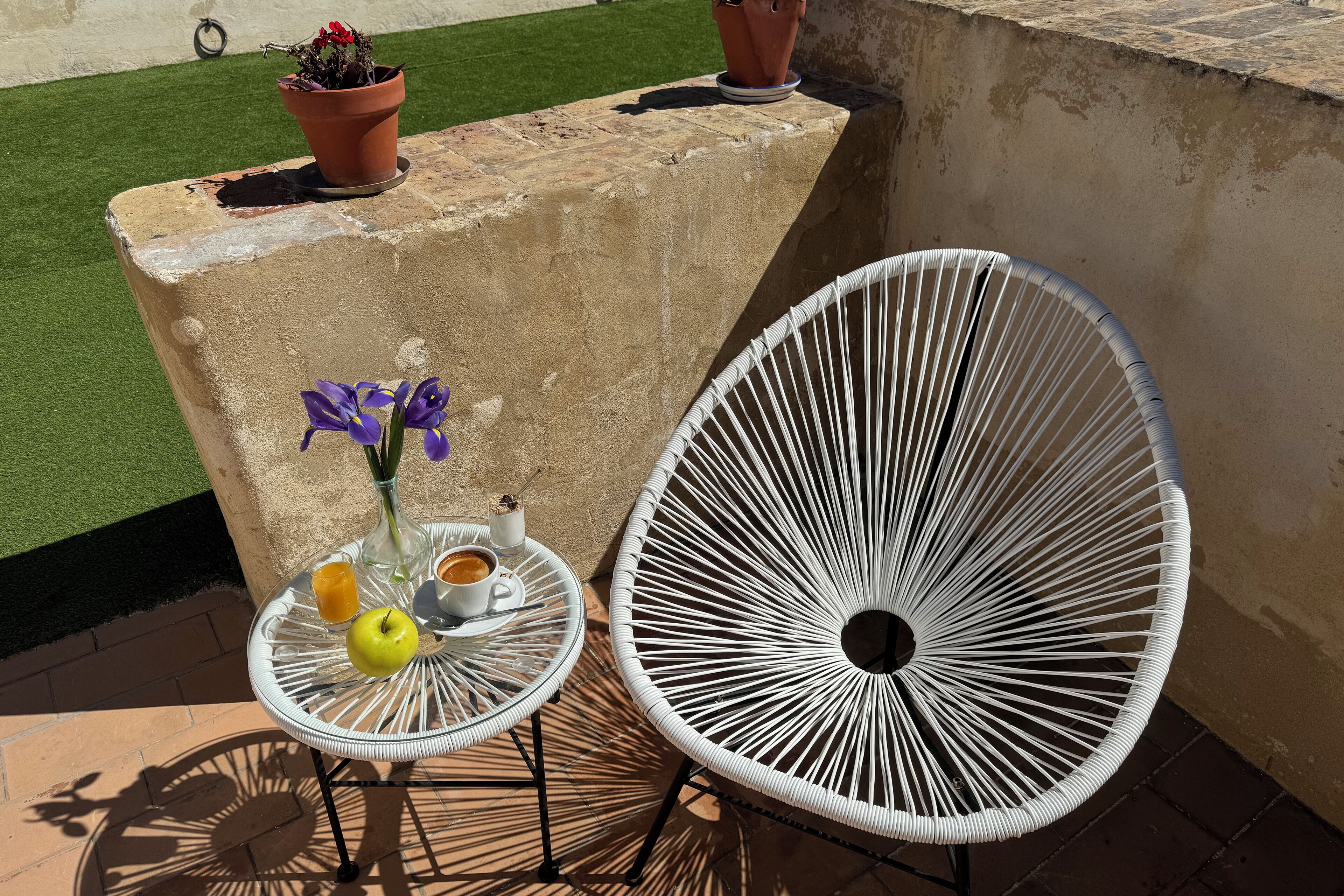A patio area with a white metal chair, a small round table with a yellow apple, a vase with purple flowers, and a terracotta pot with red flowers.