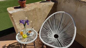 A patio area with a white metal chair, a small round table with a yellow apple, a vase with purple flowers, and a terracotta pot with red flowers.