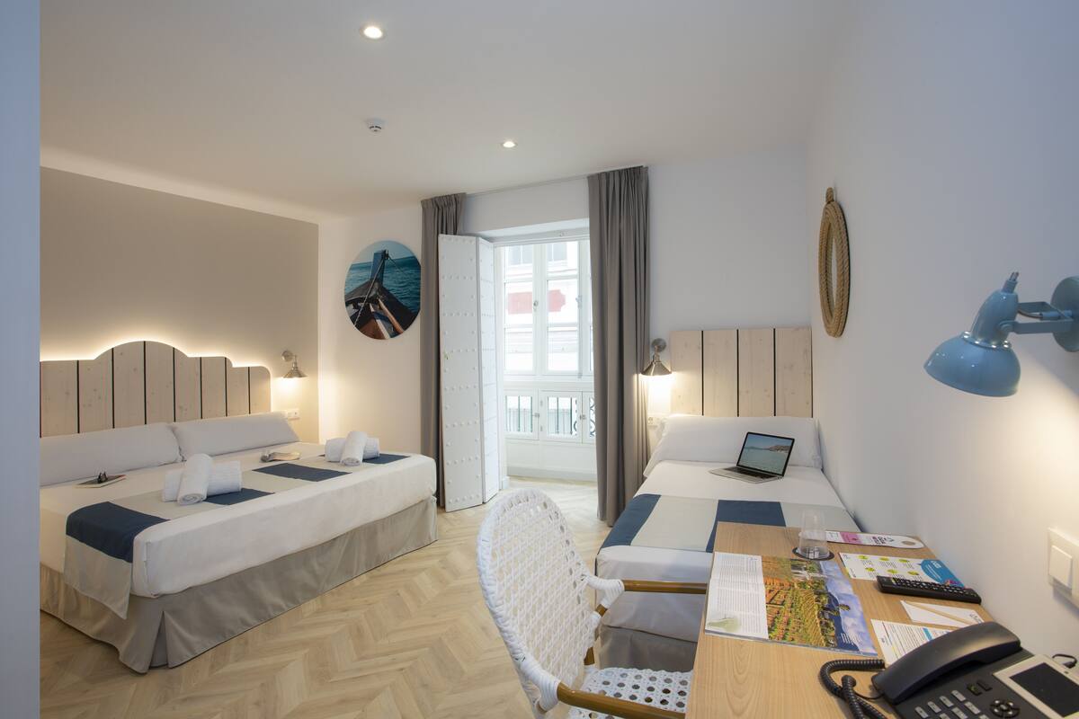 Een hotelkamer met twee bedden, een bureau met een laptop, een stoel en uitzicht op zee door een raam.