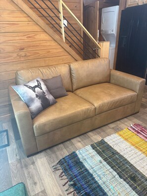 Un sofá de cuero color bronce con dos almohadas, una escalera de madera, una lavadora y un refrigerador negro.