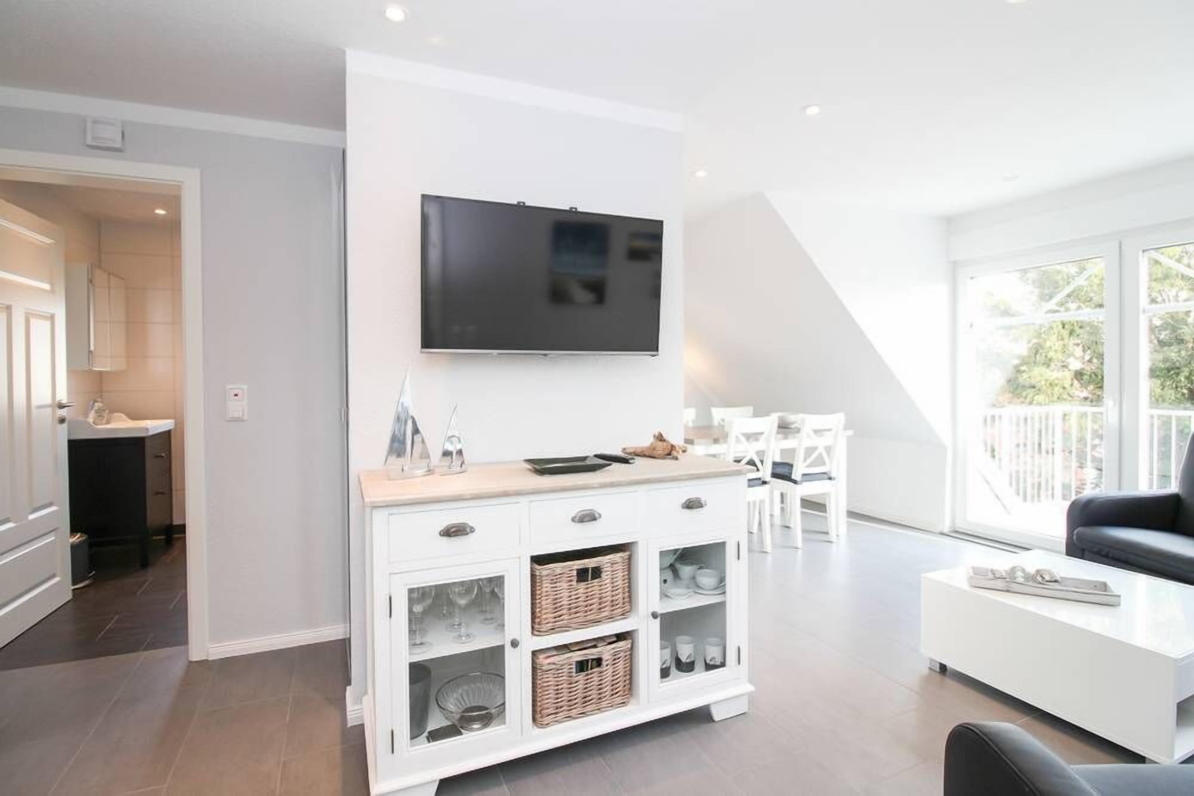 Uma sala de estar moderna com um rack branco, uma TV de tela plana e um espaço de jantar com mesa e cadeiras brancas.