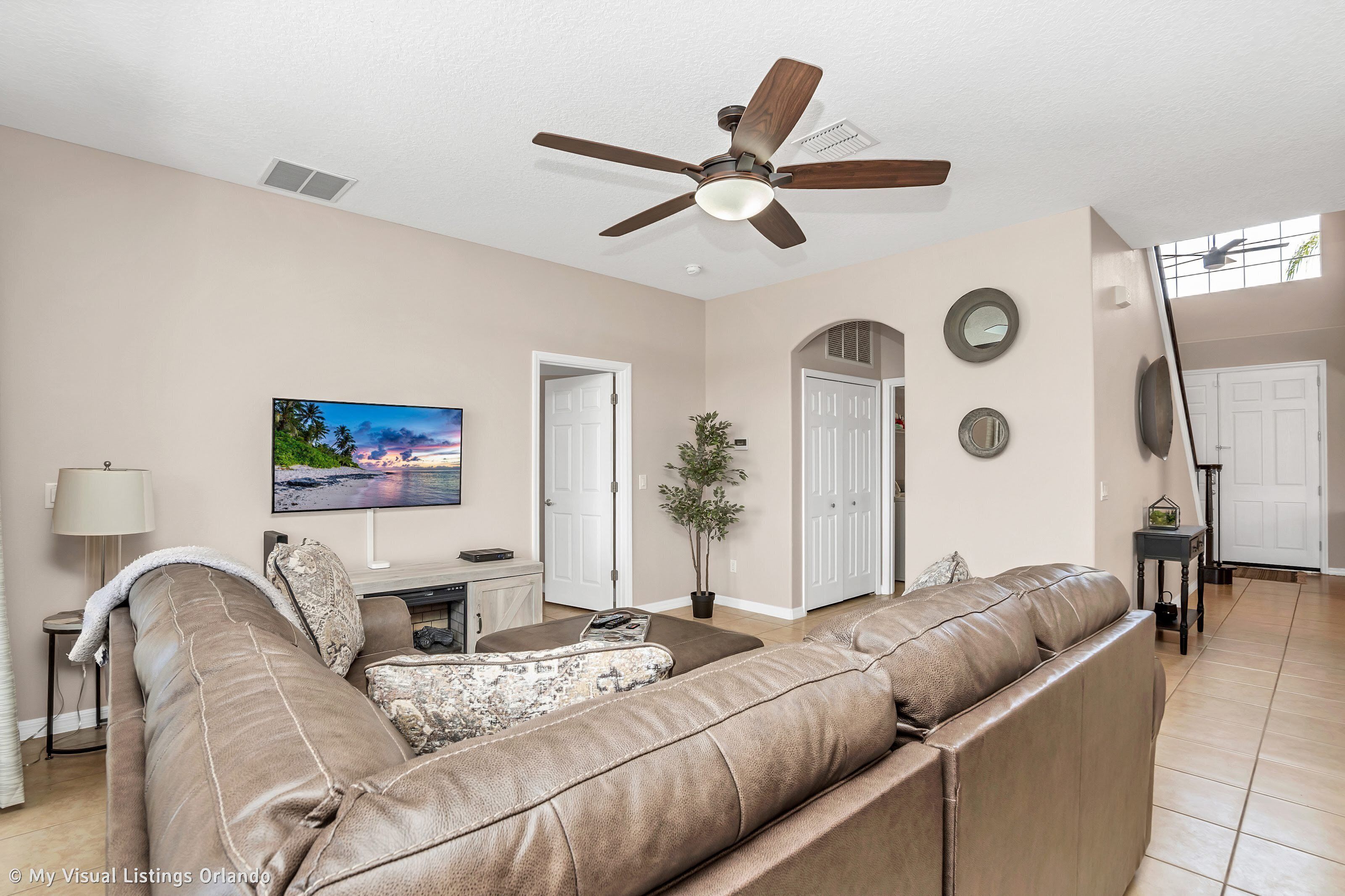 A living room with a brown leather sofa, a television, a ceiling fan, and a potted plant.