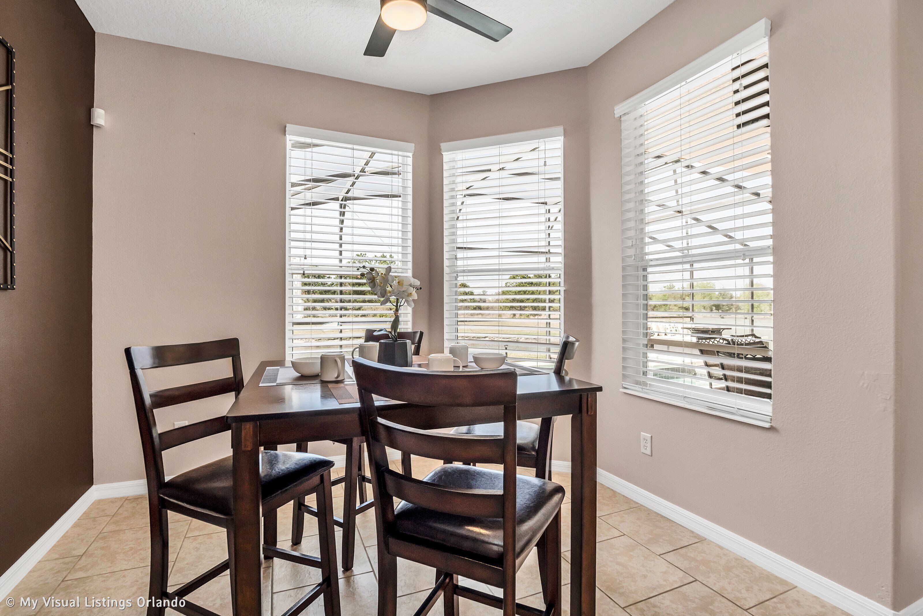 A dining area with a table and four chairs, large windows with blinds, and a ceiling fan.