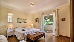 A bedroom with two beds, a bench, a nightstand with a lamp, and a window with curtains. - Villa Luz - Stunning 4-Bed Villa (Vilamoura)