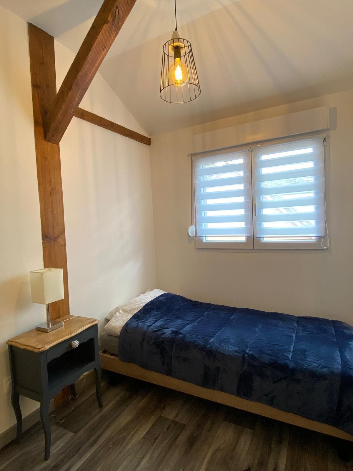Une chambre avec un lit simple, une table de chevet avec une lampe, une fenêtre avec des stores et un luminaire suspendu.
