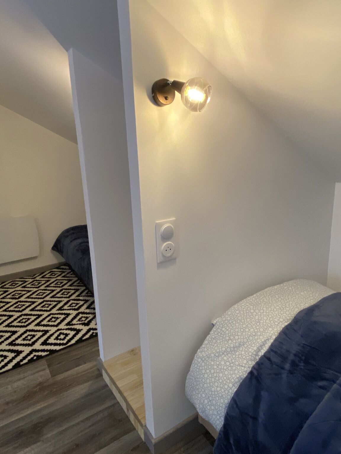 Un dormitorio con cama, un tapete estampado, una lámpara de pared y un enchufe.
