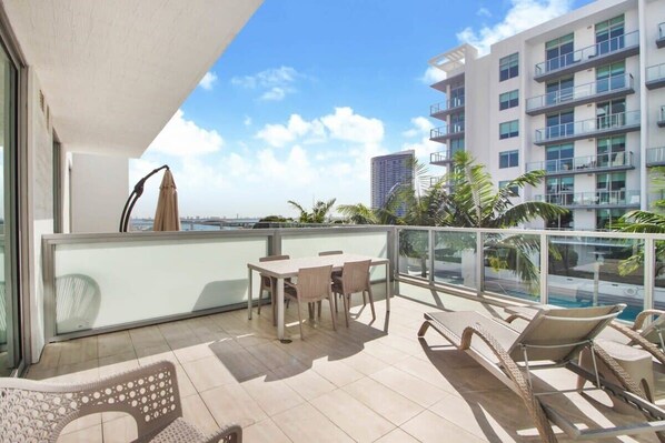 A balcony with a table and chairs, a lounge chair, and a view of a high-rise building and palm trees. - Large Studio W/Oversized Balcony & Bay Views! (Miami)