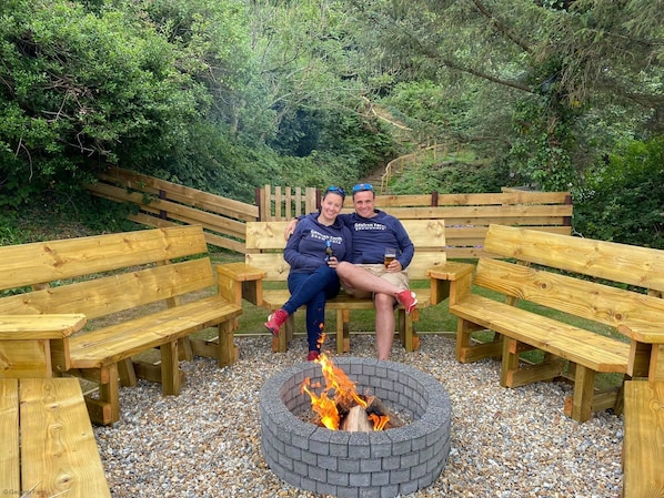 Two people sitting on wooden benches around a circular fire pit.