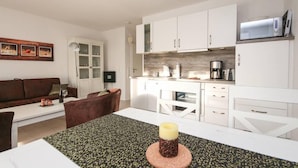 A modern kitchen with white cabinets, a microwave, and a coffee maker. There is a dining table with a candle and a patterned table runner. - Leystuuv (Krummhoern)