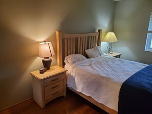 Un lit bien fait, avec une couette blanche et des oreillers, une table de chevet en bois avec une lampe, et une petite table de nuit avec une horloge.