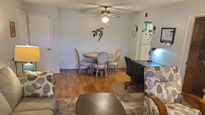 Ein Wohnzimmer mit einer Couch, einem Couchtisch, einem gemusterten Sessel, einem Esstisch mit Stühlen, einem Deckenventilator und einem Kühlschrank.
